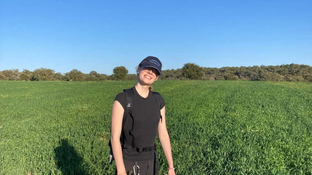 Jag står i svarta kläder ämnade för fältarbete och vandring, på ett fält med gröna grödor och blå himmel. Jag håller en karta i handen och har en GPS hängande på höften.