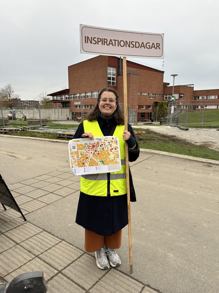 Nicole står på LTH-campus iklädd en neongul reflexväst med en karta och en skylt där det står "inspirationsdagarna".